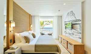 Et lyst hotellrom med en dobbeltseng, veggmontert TV og store vinduer som vender ut mot havet. (AI-generert tekst)