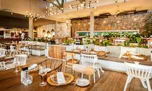 Innendørs interiør av en restaurant med trebord og stoler, murvegger, hengende lamper og grønne planter som dekorasjon. (AI-generert tekst)