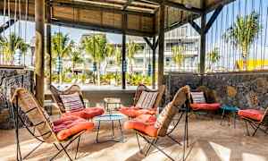 Et utendørsområde med sitteplasser, med kurvstoler og fargerike puter, samt et bord på sandgulvet. Tretak og grønne planter kan ses i bakgrunnen. (AI-generert tekst)