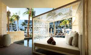 En solseng med hvite puter, plassert utendørs omgitt av hvit dekorativ struktur. I bakgrunnen er det palmer, et basseng og moderne bygninger. (AI-generert tekst)