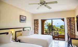 Et hotellrom med to senger, en takvifte og en balkong med utsikt mot tropiske palmer og havet. (AI-generert tekst)