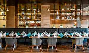 En elegant restaurant med moderne innredning. Bordene er dekket med glass og servietter, og sofaene er pyntet med puter. Hyller med dekorative gjenstander og planter er synlige i bakgrunnen. (AI-generert tekst)