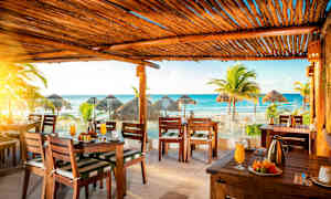 En restaurant med utsikt over stranden og havet. I forgrunnen sees dekkede bord og trestoler. I bakgrunnen er det palmer og stråtak på stranden. (AI-generert tekst)