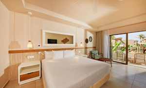 Et hotellrom med en stor seng, nattbord og en lenestol med bord. En glassdør fører ut til en balkong med utsikt over et frodig område. (AI-generert tekst)