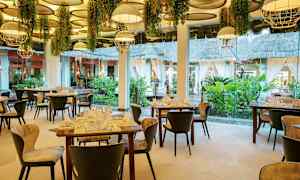 Interiør av en elegant restaurant med dekorerte bord og stoler, hengende lamper og planter, samt store vinduer med utsikt mot en frodig hage. (AI-generert tekst)