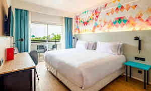 Et hotellrom med en dobbeltseng, fargerikt veggdesign, skrivebord, og balkong med to stoler. (AI-generert tekst)