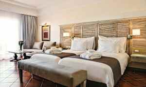 Et hotellrom med to separate senger, en sofa, et bord med en blomster vase og et vindu med gardiner. (AI-generert tekst)