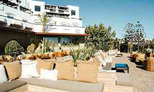 Et utendørs loungeområde med sittegrupper, puter og planter, med en moderne bygning i bakgrunnen. (AI-generert tekst)