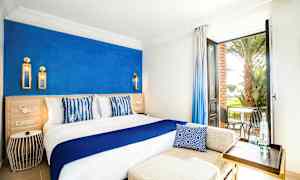 Et lyst hotellrom med en blå aksentvegg, en stor seng med hvit og blå innredning, samt en åpen dør med utsikt over en terrasse med stoler og palmer. (AI-generert tekst)