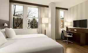 Hotellrom med to enkeltsenger, skrivebord, stol og TV, samt store vinduer med utsikt mot en bymiljø. (AI-generert tekst)