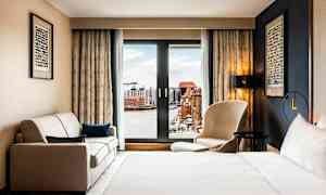 Et hotellrom med en seng, en sofa og en stol foran et stort vindu med utsikt over en by og vann. (AI-generert tekst)