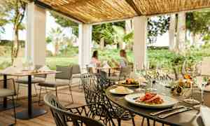 En utendørs restaurant med dekte bord og grønne planter i bakgrunnen. (AI-generert tekst)