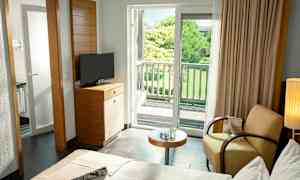 Et hotellrom med seng, lenestol, bord, TV og en balkong som vender mot en frodig hage. (AI-generert tekst)