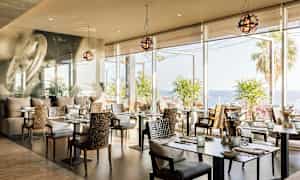 En restaurant med moderne bord og stoler, store vinduer som viser utsikt over havet og palmetrær. (AI-generert tekst)