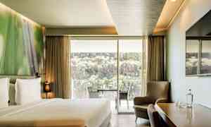 Et hotellrom med en dobbeltseng, sittegruppe, et bord med stoler samt en stor glassdør som åpner opp til en balkong med utsikt over natur og klipper. (AI-generert tekst)