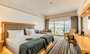 Hotellrom med to enkeltsenger, en sittegruppe og et skrivebord. Store vinduer med utsikt over havet og omgivelsene. (AI-generert tekst)