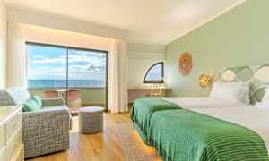 Interiør av et hotellrom med grønne detaljer, to senger, en sittegruppe og en balkong med havutsikt. (AI-generert tekst)