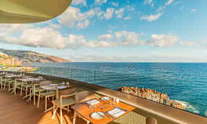 Restaurantterrasse med bord og stoler, med utsikt over havet og en klipperik kyst under en klar himmel. (AI-generert tekst)