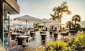 Utendørsterrasse med bord og stoler under hvite parasoller, omgitt av grønne planter og palmer. (AI-generert tekst)