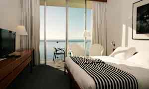 Hotellrom med seng, lenestoler og utsikt over havet gjennom store glassvinduer. (AI-generert tekst)