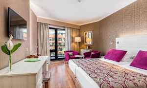 Hotellrom med dobbeltseng, skrivebord, to lilla lenestoler og balkong. (AI-generert tekst)