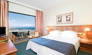 Hotellrom med dobbeltseng, nattbord og balkong med utsikt over havet. (AI-generert tekst)