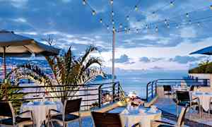 Utendørs serveringsområde med dekkede bord og stoler, omgitt av lyslenker og med utsikt over byen og havet. (AI-generert tekst)