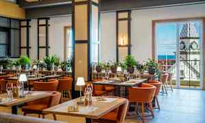 Et godt opplyst restaurantinteriør med bord, stoler og grønn plantepynt. I bakgrunnen er det store vinduer som viser en utsikt av en bygning med et klokketårn. (AI-generert tekst)