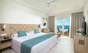 Et hotellrom med dobbeltseng, sittegruppe og balkong med havutsikt. (AI-generert tekst)