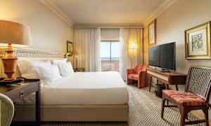 Et hotellrom med en stor seng, nattbord, sofa, TV og et skrivebord. Gardiner og glassdører fører til en balkong. (AI-generert tekst)