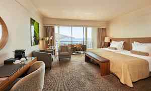 Hotellrom med dobbeltseng, sittegruppe og balkong med utsikt over havet. (AI-generert tekst)