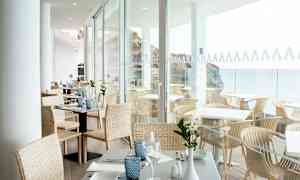 Lyst og åpent spiseområde med dekkede bord. Utsikt mot havet gjennom glassvegger. (AI-generert tekst)