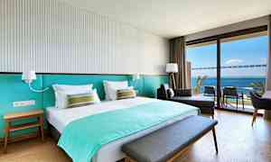 Et hotellrom med en dobbeltseng, en sittemøbel og en balkong med utsikt over havet. (AI-generert tekst)