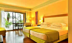 Et hotellrom med to enkeltsenger, grønne laken og en sittegruppe, med utsikt over utendørs palmer gjennom store glassvinduer. (AI-generert tekst)