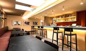 Et moderne barområde med hovedsakelig svarte bord og stoler samt en godt opplyst bardisk fylt med flasker og glass. (AI-generert tekst)