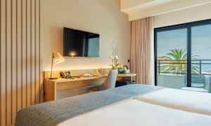 Et hotellrom med skrivebord, stol og TV, samt en balkong med utsikt over palmer og havet. (AI-generert tekst)