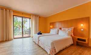 Et hotellrom med to senger dekket med hvitt sengetøy, et trehodegjerde og en balkong med utsikt. Rommet har gulmalte vegger og tregulv. (AI-generert tekst)