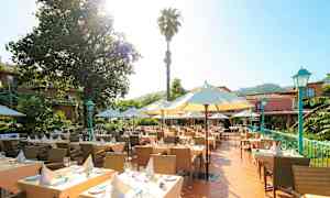 En utendørs terrasse med dekkede bord omgitt av parasoller og grøntområder. I bakgrunnen er det en bygning omgitt av trær. (AI-generert tekst)