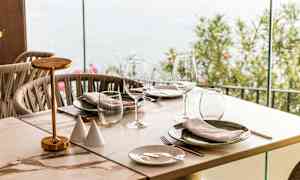 Et dekket bord med glass og tallerkener, ved siden av en glassvegg som vender utendørs. (AI-generert tekst)