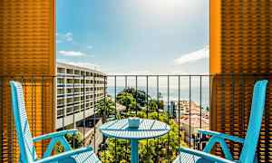 To blå stoler og et bord på en balkong med utsikt over havet og omkringliggende bygninger. (AI-generert tekst)