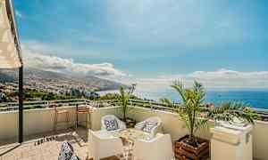 Utsikt fra en terrasse med sittemøbler, palmer og panoramautsikt over by, fjell og hav. (AI-generert tekst)
