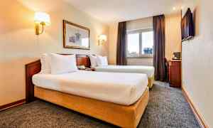 Bilde av et hotellrom med to enkeltsenger, et vindu med gardiner, en veggdekorasjon og et skrivebord med en stol. (AI-generert tekst)