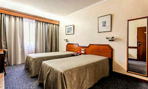 Bilde av et hotellrom med to enkeltsenger, et skrivebord og et vindu med gardiner. (AI-generert tekst)