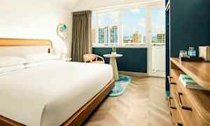 Et hotellrom med en stor seng, et vindu med utsikt over en by med høyhus og himmel, samt et møbel med vannflasker og bøker. (AI-generert tekst)