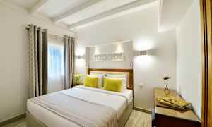 Et lyst soverom med en dobbeltseng, dekorert med to gule puter og et sengeteppe. Vinduet har gardiner, og det er en arbeidsplass med skrivebord og lampe. (AI-generert tekst)