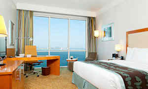 Et moderne hotellrom med en stor seng, skrivebord og utsikt gjennom store vinduer mot havet og en by i bakgrunnen. (AI-generert tekst)