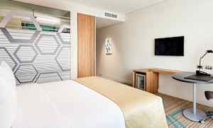 Et hotellrom med en seng dekket i hvite og beige farger. Rommet inkluderer også et skrivebord, en TV på veggen og et minikjøleskap. (AI-generert tekst)