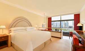 Hotellrom med dobbeltseng, nattbord og utsikt gjennom store vinduer over en bysilhuett. (AI-generert tekst)