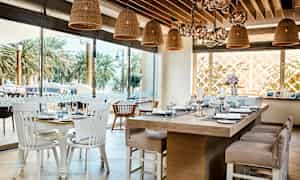 Interiør av en restaurant med bord dekket med servise og glass, samt rottinglamper som henger fra taket. Utenfor er det palmer. (AI-generert tekst)