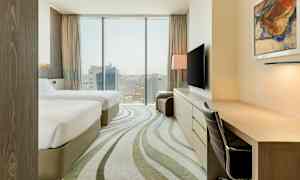 Et hotellrom med to senger, skrivebord, stol og TV. Vindu med utsikt over en by. (AI-generert tekst)